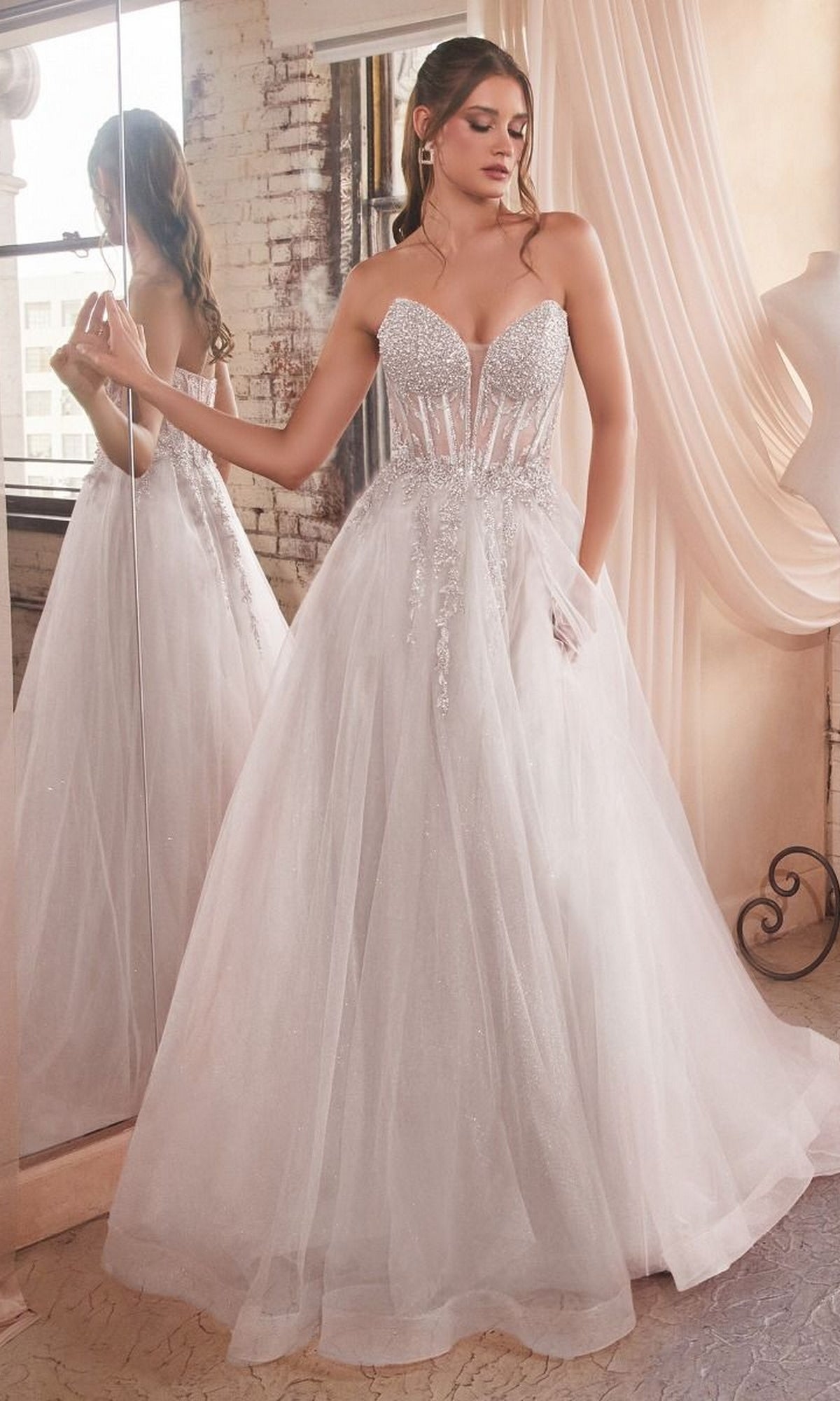 A woman stands indoors next to a large mirror and exposed brick wall, wearing the Ladivine CD0230W Strapless Long White Bridal Gown with a sparkly tulle skirt and embellished bodice.